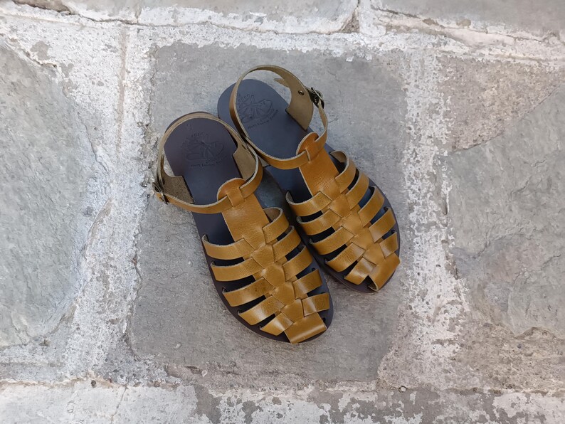 May include: A pair of mustard yellow leather sandals featuring a woven cage design, a closed toe, and an adjustable ankle strap with a metal buckle. The footbed is dark brown with a visible embossed logo stamp near the heel area of each sandal.