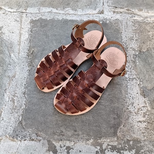 May include: A pair of dark brown leather fisherman sandals featuring a woven strap design, an adjustable ankle strap with a gold-tone metal buckle, and a light tan leather footbed with a stamped logo and the number 7 visible on the heel area.