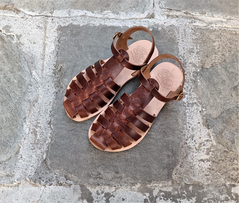 May include: A pair of dark brown leather fisherman sandals featuring a woven strap design, an adjustable ankle strap with a gold-tone metal buckle, and a light tan leather footbed with a stamped logo and the number 7 visible on the heel area.