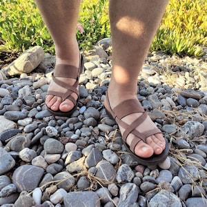 May include: A pair of brown leather sandals with criss-cross straps. The sandals are being worn by someone standing on a bed of small, smooth stones.