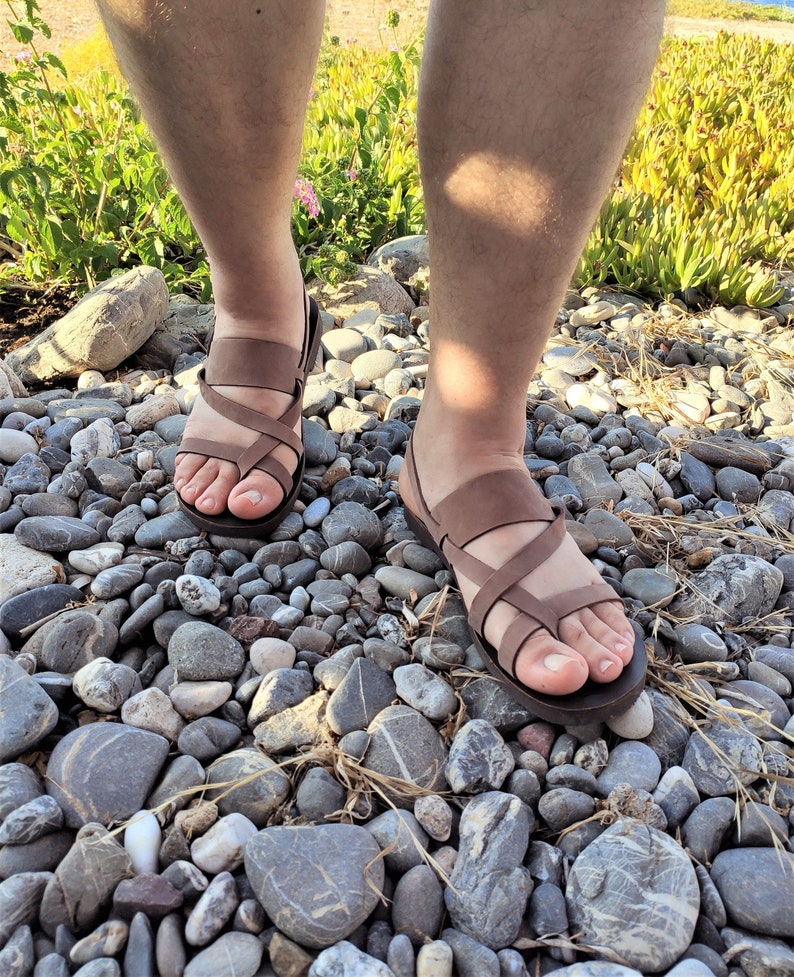 May include: A pair of brown leather sandals with criss-cross straps. The sandals are being worn by someone standing on a bed of small, smooth stones.