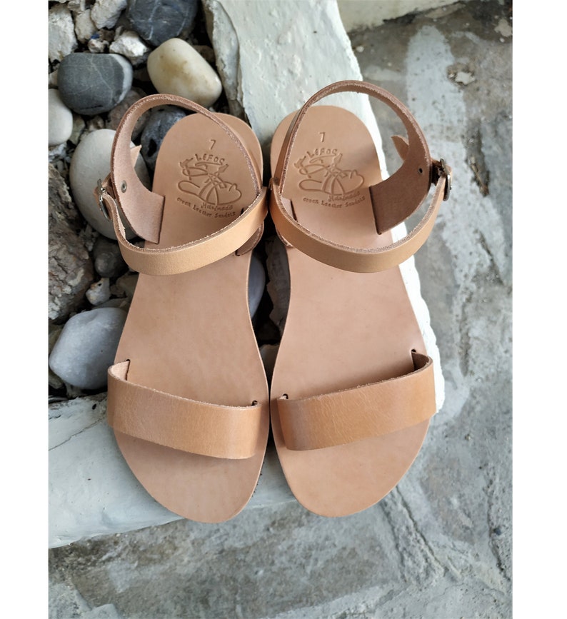 K&ouml;nnte beinhalten: Ein Paar hellbraune Ledersandalen mit zwei Riemen. Die Sandalen haben einen Schnallenverschluss und der Markenname "LIFOS" ist auf den Riemen gepr&auml;gt.