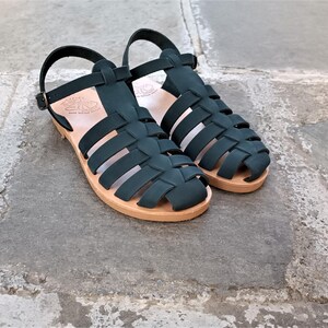 May include: A pair of dark green fisherman sandals featuring a woven strap design, an adjustable ankle strap with a metal buckle, and a light tan flat sole. The interior of the sole features a stamped logo and text detail.