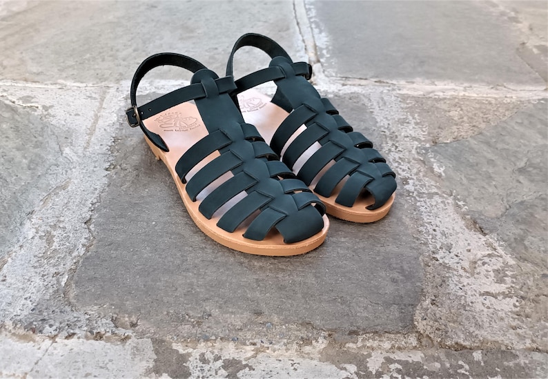 May include: A pair of dark green fisherman sandals featuring a woven strap design, an adjustable ankle strap with a metal buckle, and a light tan flat sole. The interior of the sole features a stamped logo and text detail.