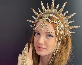 May include: A woman with long red hair wearing a gold and crystal crown with dangling chains. She is also wearing a gold and crystal bracelet on her wrist and a gold and crystal ring on her finger. The background is a dark gray.