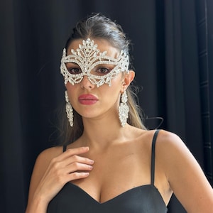 Könnte beinhalten: Eine silberne Strass-Maske mit floralem Design, kombiniert mit passenden Chandelier-Ohrringen. Die Maske bedeckt die Augen und die Stirn. Die Frau trägt ein schwarzes Kleid.