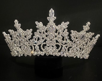 May include: A silver crown with a large, intricate design made of clear rhinestones. The crown is on a black background.