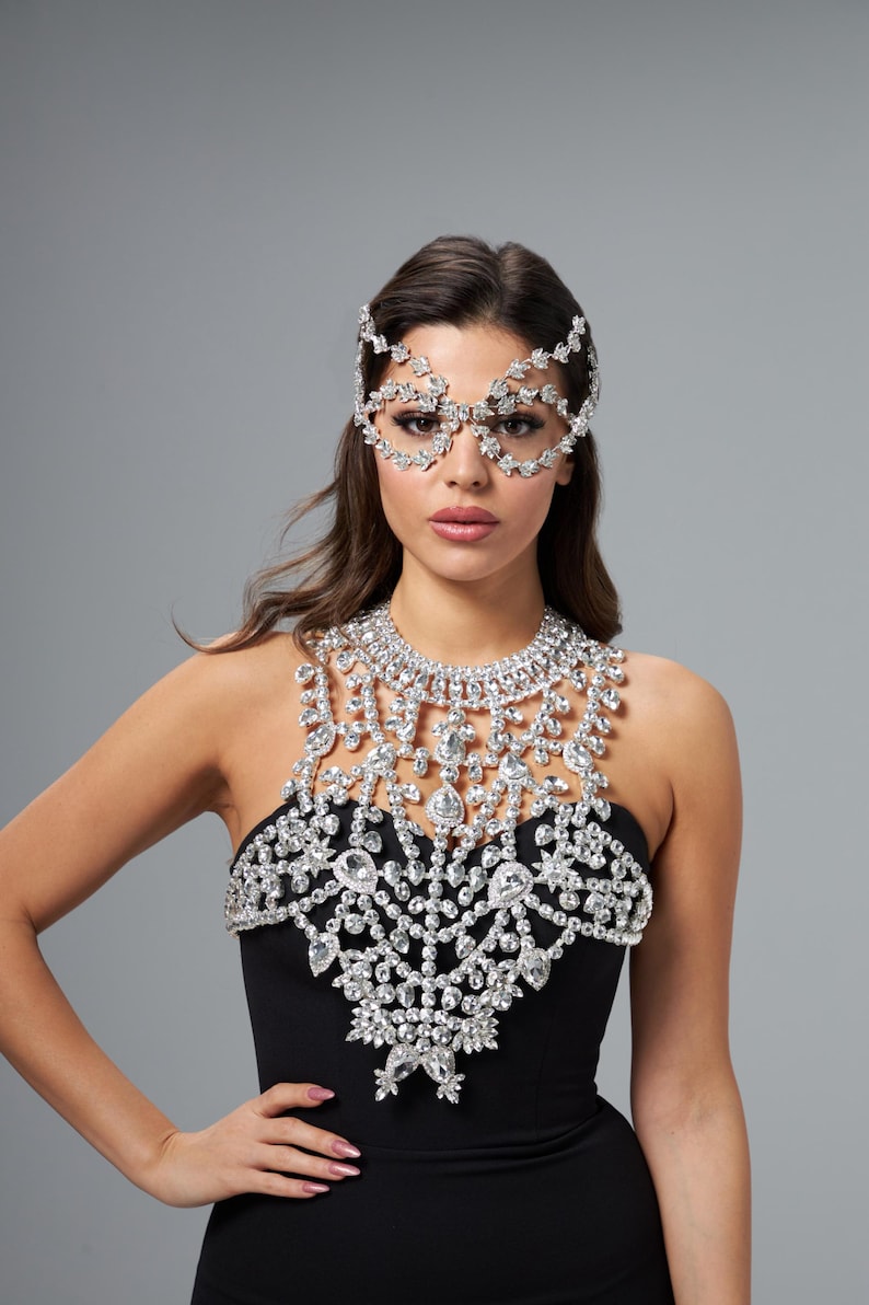May include: A woman in a black dress, adorned with a dazzling crystal necklace, headpiece, and eye mask. The intricate jewellery is made of sparkling crystals, creating a glamorous and elegant look.