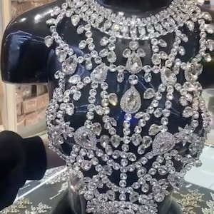 May include: A glamorous, bejeweled bodice and necklace displayed on a black mannequin. The jewelry is covered in numerous clear, sparkling crystals of different sizes, creating a luxurious design. The crystals are arranged in an intricate, interconnected pattern.