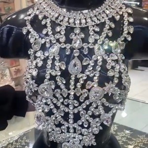 May include: A glamorous, silver-coloured bodice displayed on a black mannequin. The intricate design features numerous sparkling, clear gemstones of various shapes and sizes, creating a luxurious effect. The piece is displayed in a shop setting, suggesting formal wear or special occasion attire.