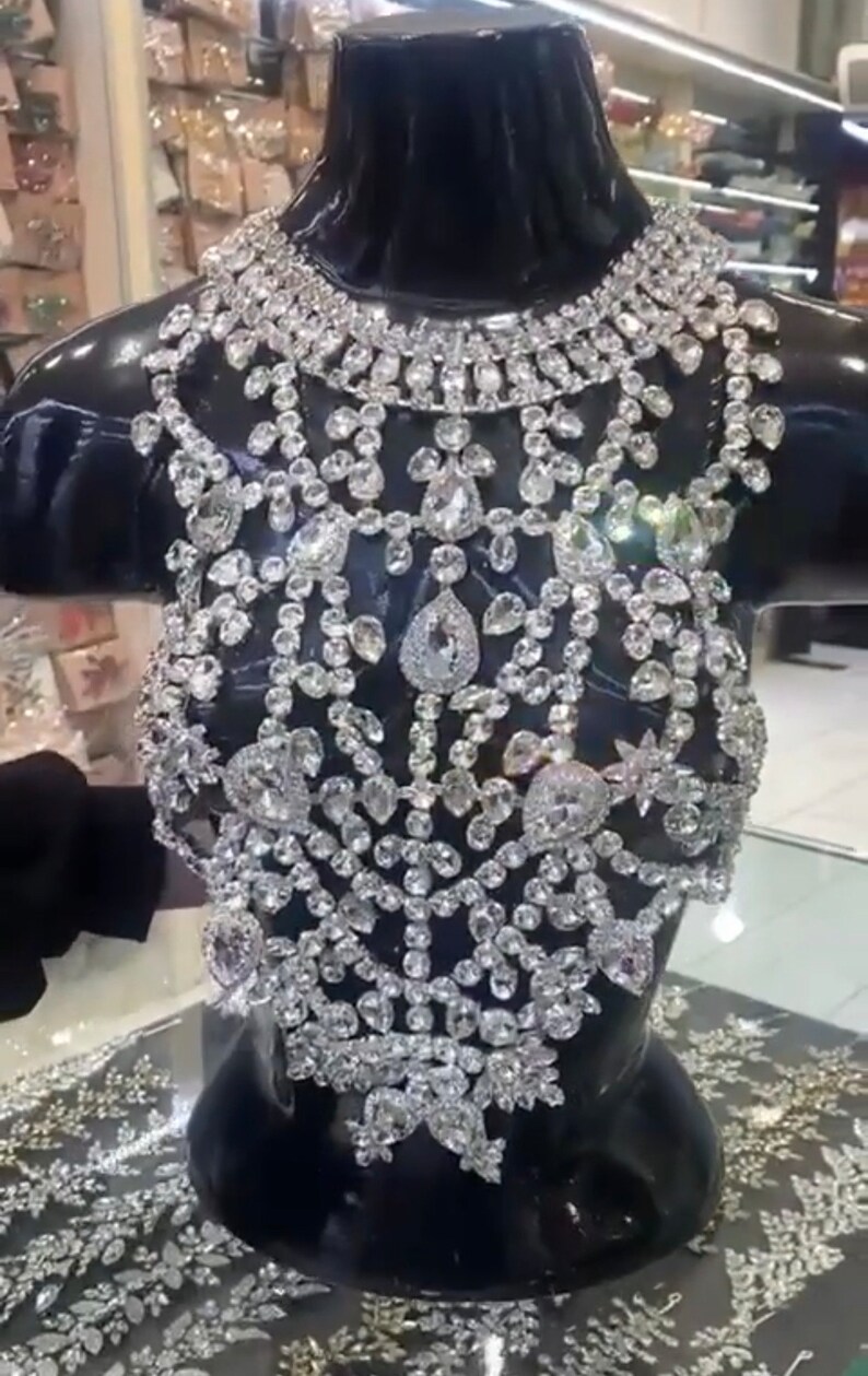 May include: A glamorous, silver-coloured bodice displayed on a black mannequin. The intricate design features numerous sparkling, clear gemstones of various shapes and sizes, creating a luxurious effect. The piece is displayed in a shop setting, suggesting formal wear or special occasion attire.