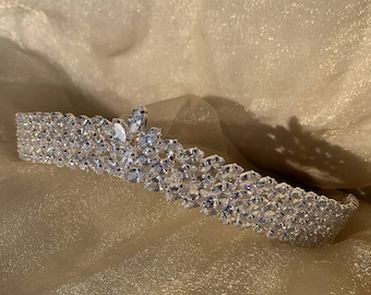 May include: A silver tiara with a row of sparkling clear rhinestones. The tiara is curved and has a pointed top.