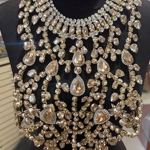 May include: A striking gold-coloured statement necklace with detailed craftsmanship. The necklace is adorned with numerous clear and champagne-hued crystals of varying sizes, arranged in a floral and geometric design. It features teardrop and round-cut stones, creating a luxurious accessory.