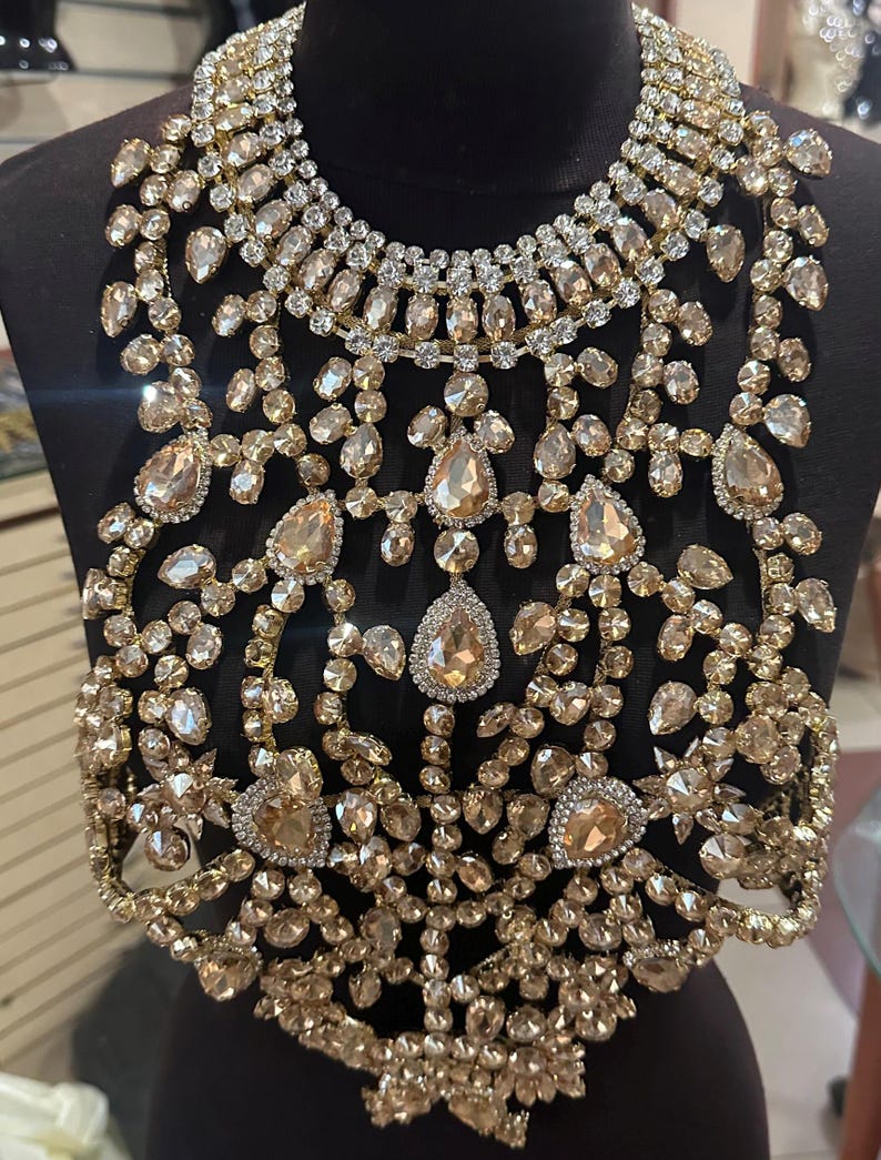 May include: A striking gold-coloured statement necklace with detailed craftsmanship. The necklace is adorned with numerous clear and champagne-hued crystals of varying sizes, arranged in a floral and geometric design. It features teardrop and round-cut stones, creating a luxurious accessory.