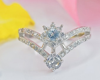 May include: A silver ring with a V-shaped band and a center stone surrounded by smaller stones. The center stone is a light blue gemstone, and the smaller stones are clear.
