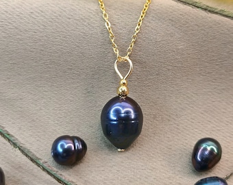 May include: A gold chain necklace with a single dark blue pearl pendant. The pearl is teardrop shaped and has a slight iridescent sheen. There are three additional dark blue pearls on the surface below the necklace.