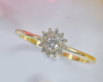 May include: A gold ring with a round, clear stone surrounded by smaller stones. The ring is on a white surface with pink and yellow fabric in the background.