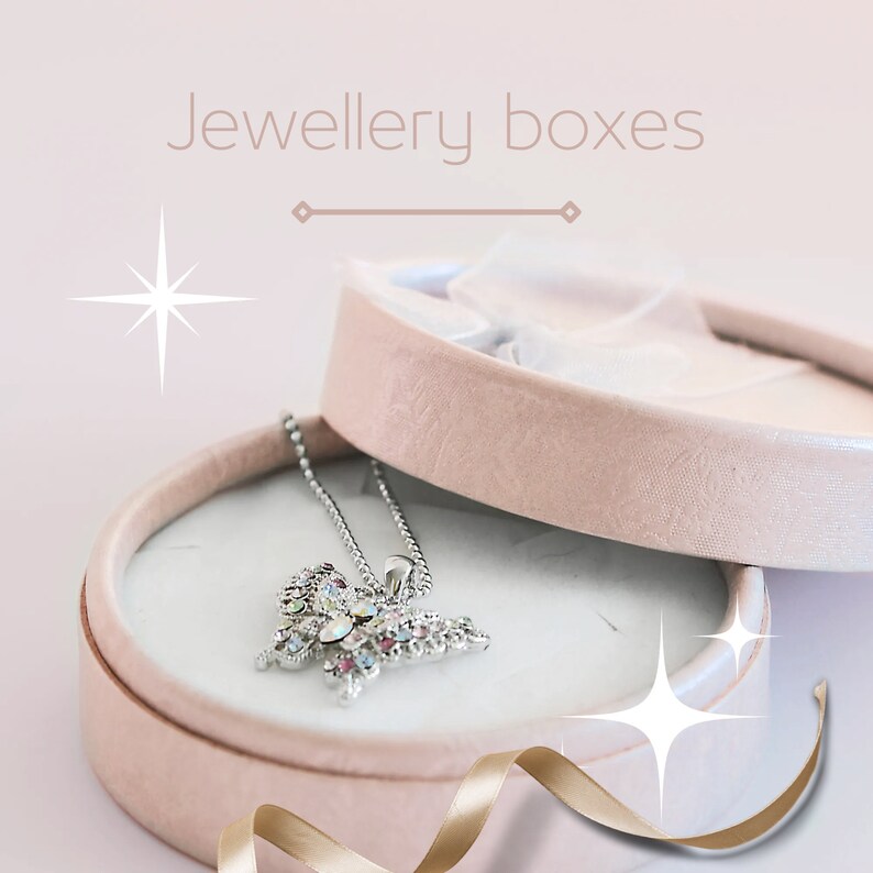 May include: A pink round jewelry box with a white satin interior. The box is open and a silver butterfly necklace with colorful rhinestones is inside. The box is on a white surface with a gold ribbon.