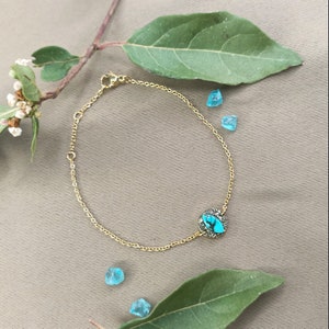 May include: A gold chain bracelet with a turquoise stone set in a circular bezel. The stone is surrounded by smaller stones. The bracelet is on a brown surface with green leaves and blue stones.