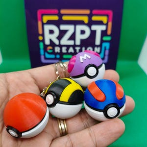 Può includere: Quattro portachiavi Poké Ball colorati stampati in 3D in rosso, giallo, blu e viola, ciascuno con una base bianca e dettagli neri. I portachiavi sono tenuti in mano, con uno sfondo viola che recita "RZPT CREATION".