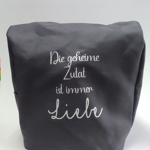 Op de afbeelding: Een donkergrijze afdekking voor keukenapparatuur met de tekst "Die geheime Zutat ist immer Liebe" in witte letters. De hoes is gemaakt van een gladde stof en is ontworpen om over een keukenapparaat te passen.