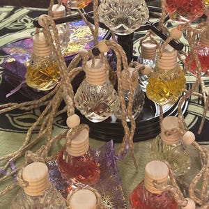 Peut inclure: Une collection de petits flacons en verre à facettes remplis de liquides colorés, surmontés d'un bouchon en bois clair et suspendus par une cordelette tressée. Les flacons sont disposés sur un présentoir en métal noir, avec des teintes transparentes, rouges et jaunes.