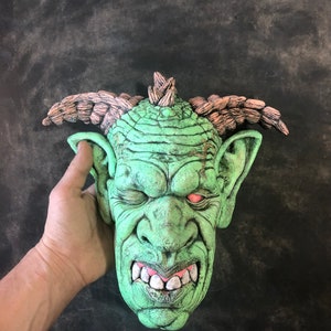May include: A green goblin mask with a spiky mohawk, brown horns, and a menacing expression. The mask has a textured surface and a red eye.
