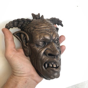 May include: A bronze sculpture of a grotesque face with a spiky mohawk, large ears, and a snarling expression.