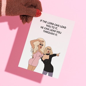 May include: A greeting card with the text "IF THE LORD CAN LEAD YOU TO IT, HE CAN LEAD YOU THROUGH IT." features two cartoon women. One is in a pink dress, the other in a black dress. The card is held by a person wearing a brown glove.