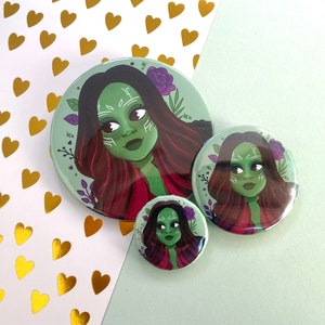 May include: Three round green, red and purple buttons featuring a cartoon image of a woman with green skin and red hair. The woman is wearing a red and black shirt.