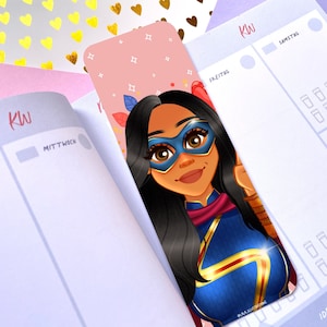 Puede incluir: Un marcapáginas rosa con una ilustración de dibujos animados de un superhéroe que lleva un traje azul y dorado con una capa roja. El superhéroe tiene piel morena, cabello negro y una máscara de ojos azul. El fondo es rosa con estrellas blancas y purpurina dorada.