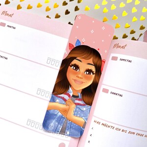 Puede incluir: Una agenda rosa y blanca con un marcapáginas que presenta una ilustración de dibujos animados de una mujer con cabello castaño y una chaqueta vaquera. El marcapáginas tiene un fondo rosa con estrellas blancas y el texto "Monat" en la parte superior.