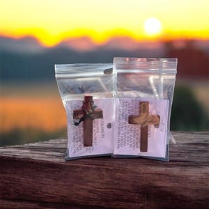 Könnte beinhalten: Zwei Holz-Kreuz-Anhänger in transparenten Plastiktüten, auf einer Holzoberfläche. Die Kreuze sind braun, eines mit einem kleinen Band. Der Hintergrund zeigt einen verschwommenen Sonnenuntergang mit Orange- und Gelbtönen.