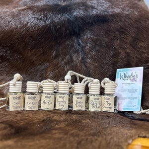 May include: A collection of small glass bottles with wooden lids, each labeled with a different scent name, including "Sweet Tobacco" and "New Boot Goofin". A card with the text "Wheelie's Whiffs" is also visible. The bottles are arranged on a brown surface.