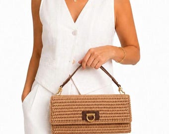 Puede incluir: Un bolso de mano rectangular de paja trenzada en color beige, con un asa de cuero marrón oscuro y un cierre de solapa de cuero a juego con un broche de metal dorado en forma de herradura. El bolso tiene una estructura horizontal.
