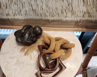 Puede incluir: Una escultura marrón oscuro de una persona en posición fetal se encuentra sobre una mesa blanca con un patrón de espiga. Un nudo de cuerda decorativo y formas geométricas de madera también están sobre la mesa. Una borla beige cuelga de las formas de madera.