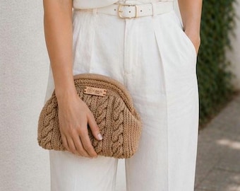 Puede incluir: Un bolso de mano tejido de color marrón claro con un diseño de punto trenzado. El bolso tiene una pequeña etiqueta rectangular de cuero con un logotipo. La persona lleva pantalones blancos y un cinturón blanco con hebilla dorada.