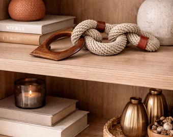 Puede incluir: Una estantería de madera exhibe objetos decorativos. Un nudo de cuerda trenzada con detalles de cuero se encuentra cerca de un asa de madera. Otros elementos incluyen jarrones, libros, una vela y pequeños objetos decorativos dorados. La estantería es de madera clara.