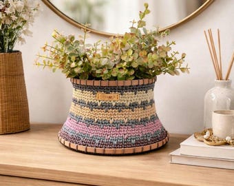 Puede incluir: Una cesta de ganchillo multicolor con ribete de cuero, llena de vegetación. La cesta presenta rayas horizontales en tonos rosa, amarillo, gris y azul. Una pequeña etiqueta está adherida al frente. La cesta está sobre una superficie de madera.