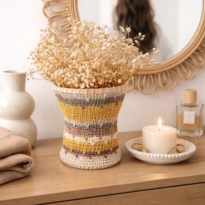 Puede incluir: Un jarrón decorativo con un diseño tejido en tonos beige, amarillo, gris y marrón, lleno de flores secas. Una vela encendida está sobre un plato blanco pequeño, y un espejo redondo está en el fondo.