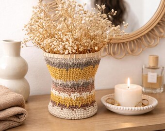 Puede incluir: Un jarrón decorativo con un diseño tejido en tonos beige, amarillo, gris y marrón, lleno de flores secas. Una vela encendida está sobre un plato blanco pequeño, y un espejo redondo está en el fondo.