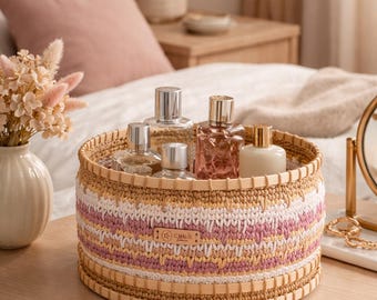 Puede incluir: Una cesta redonda tejida con un patrón crema, rosa y beige contiene varios frascos de perfume. La cesta está colocada sobre una superficie de madera clara. La cesta tiene un borde de madera en la parte superior e inferior.