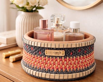 Puede incluir: Una cesta redonda de ganchillo hecha a mano con borde y base de madera, que contiene varios frascos de perfume. La cesta presenta un patrón geométrico gris, naranja y rosa. La etiqueta dice "C. Malis Design".