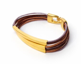 Puede incluir: Pulsera de múltiples hilos con cordones de cuero marrón y morado. Cuenta con un cierre dorado y una barra rectangular dorada. Esta pulsera es un accesorio de moda.