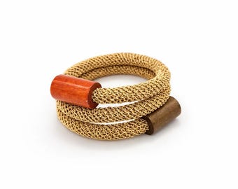 Puede incluir: Una pulsera dorada tejida con dos acentos cilíndricos de madera. Uno es de color marrón rojizo, el otro es marrón más oscuro. La pulsera está enrollada y descansa sobre un fondo blanco.