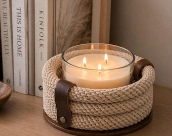 Puede incluir: Vela de tres mechas en un recipiente de vidrio transparente, colocada en un soporte de cuerda beige tejido con detalles de cuero marrón. La vela se apoya sobre una base de madera redonda.