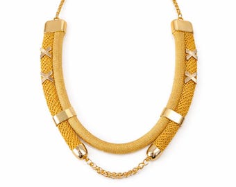 Puede incluir: Collar dorado con diseño en capas. La capa superior presenta un tubo de malla tejida, acentuado con detalles dorados en forma de X. La capa inferior es una banda dorada lisa y sólida, conectada por una delicada cadena.