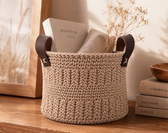 Puede incluir: Una cesta de almacenamiento redonda de ganchillo en color beige con un patrón de punto trenzado texturizado y dos asas de cuero marrón oscuro sujetas con costuras blancas. La cesta contiene un libro titulado KINFOLK TABLE y tallos.