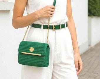 Puede incluir: Un bolso verde cruzado con una correa de cadena dorada y un cinturón verde a juego con una hebilla dorada. El bolso tiene un cierre dorado y un emblema dorado circular en la parte delantera. El bolso es de material tejido.