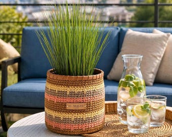 Puede incluir: Una maceta de ganchillo a rayas con ribete de cuero, que contiene hierba verde alta. La maceta tiene rayas horizontales en tonos marrón, amarillo y rosa. Una jarra de cristal y dos vasos de agua con rodajas de limón están en una bandeja redonda.